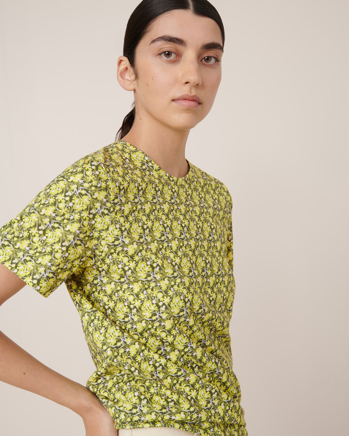 Person with long dark hair wears the Kowtow Flora Tee – Flora, a yellow floral short-sleeve shirt, paired with light pants against a plain background—ideal for displaying the fit and details of this standout top.