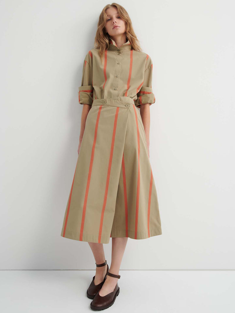A woman stands against a white wall wearing a tan shirt dress with orange vertical stripes, rolled sleeves, brown ankle-strap shoes, and the Fold Skirt – Nasturtium Stripe styled underneath for added dimension.