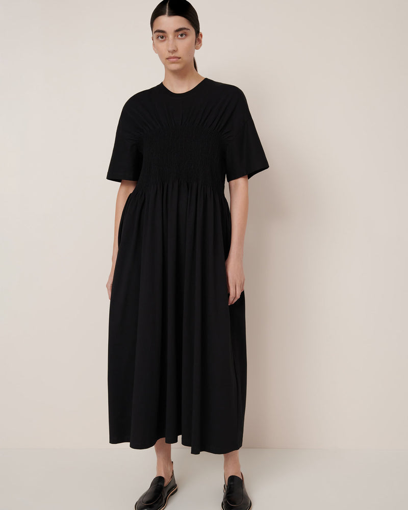 A person stands against a plain light background wearing Kowtow’s Gathered Dress – Black, an ankle-length, short-sleeved dress with gathered detailing and a relaxed fit, paired with black slip-on shoes.