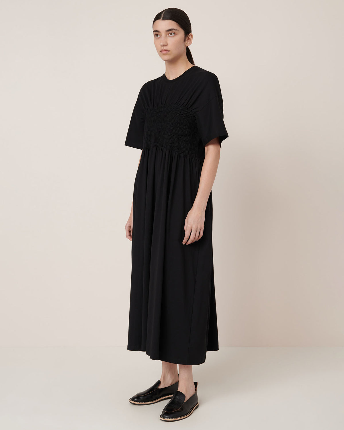A woman stands in a plain room wearing the Kowtow Gathered Dress in black, featuring short sleeves, ankle length, and a relaxed fit, paired with black slip-on shoes.