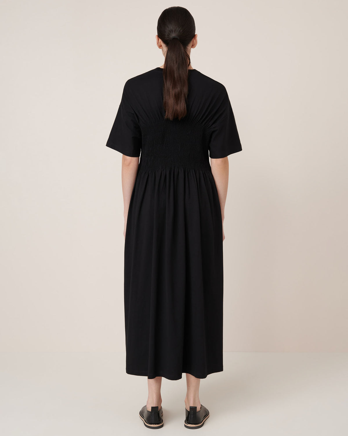 A woman faces away, wearing the Kowtow Gathered Dress – Black, a relaxed fit, ankle-length dress with short sleeves and gathered waist detailing, paired with black slip-on shoes. Garment measurements available upon request.