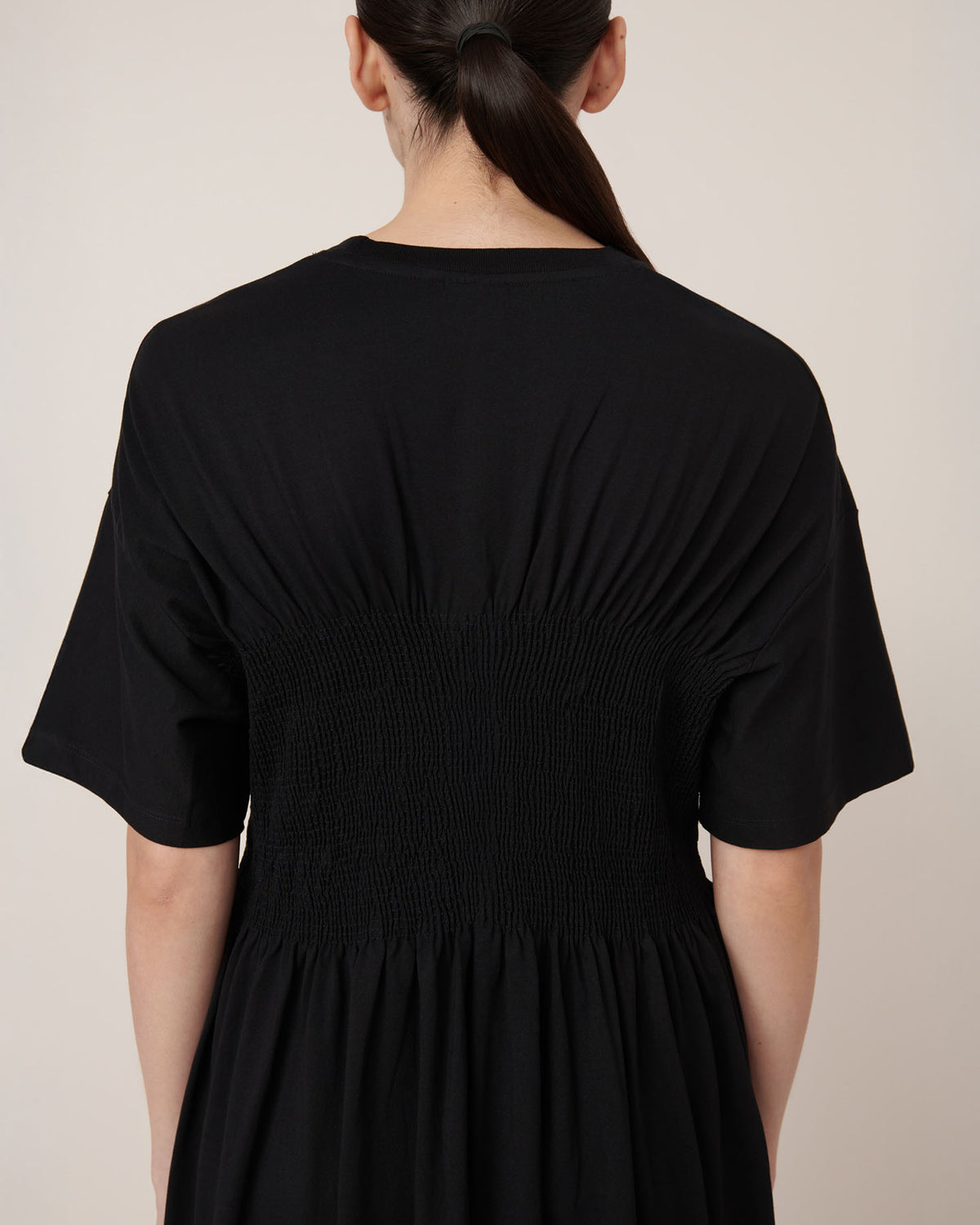 A person with long dark hair in a low ponytail is shown from the back, wearing Kowtow's Gathered Dress in black—a relaxed-fit short-sleeve style with ruched waist detailing for an effortless look.