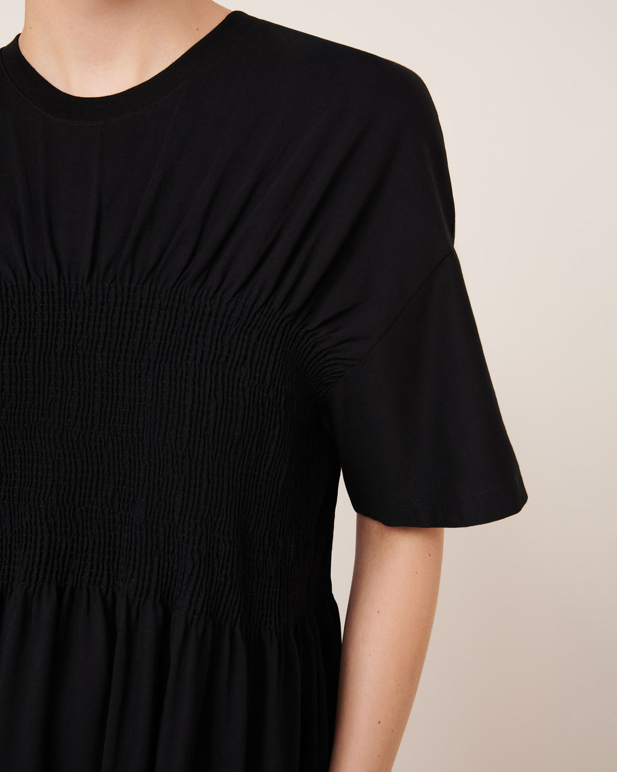 A person wears the Kowtow Gathered Dress in Black, featuring short sleeves and ruched detailing at the chest and waist, highlighting its relaxed fit in a photo from shoulders to mid-torso.