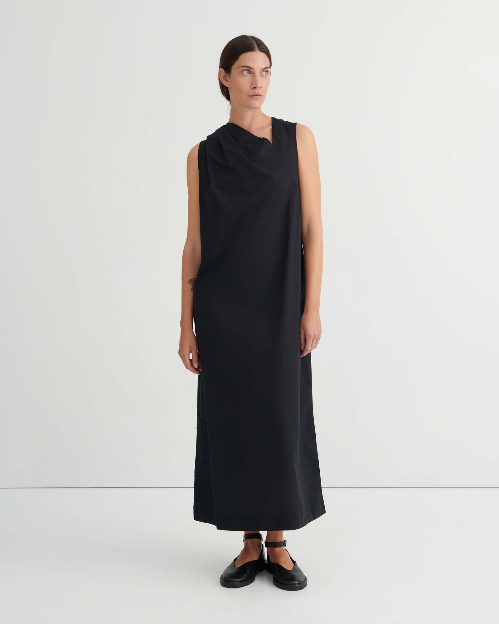 Woman stands against a white wall wearing a sleeveless, ankle-length black dress with a tied waist and black shoes.