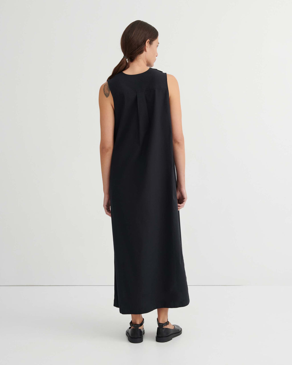 A person with long brown hair wearing a sleeveless black ankle-length dress and black sandals stands facing a white wall.
