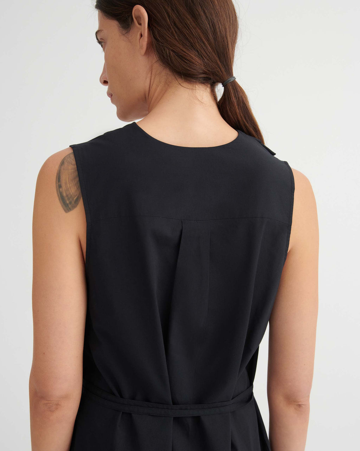 Woman with brown hair in a ponytail, wearing a sleeveless black top, is shown from the back with a visible shoulder tattoo.