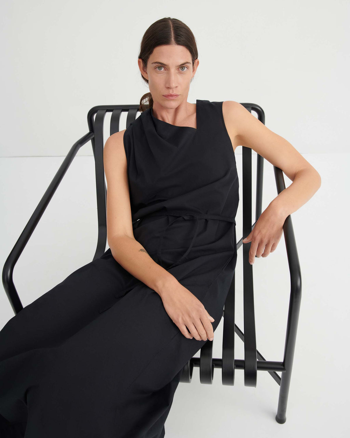 A person with long dark hair, wearing a sleeveless black dress, sits on a modern black metal chair against a plain white background.