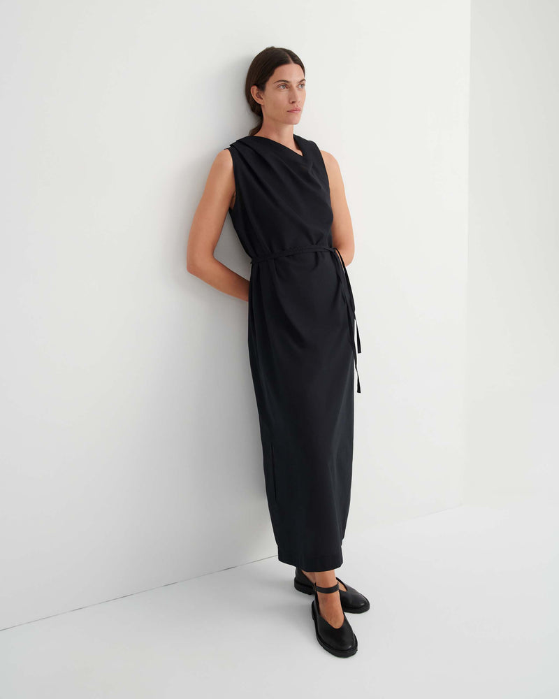 Woman stands against a white wall wearing a sleeveless, ankle-length black dress with a tied waist and black shoes.
