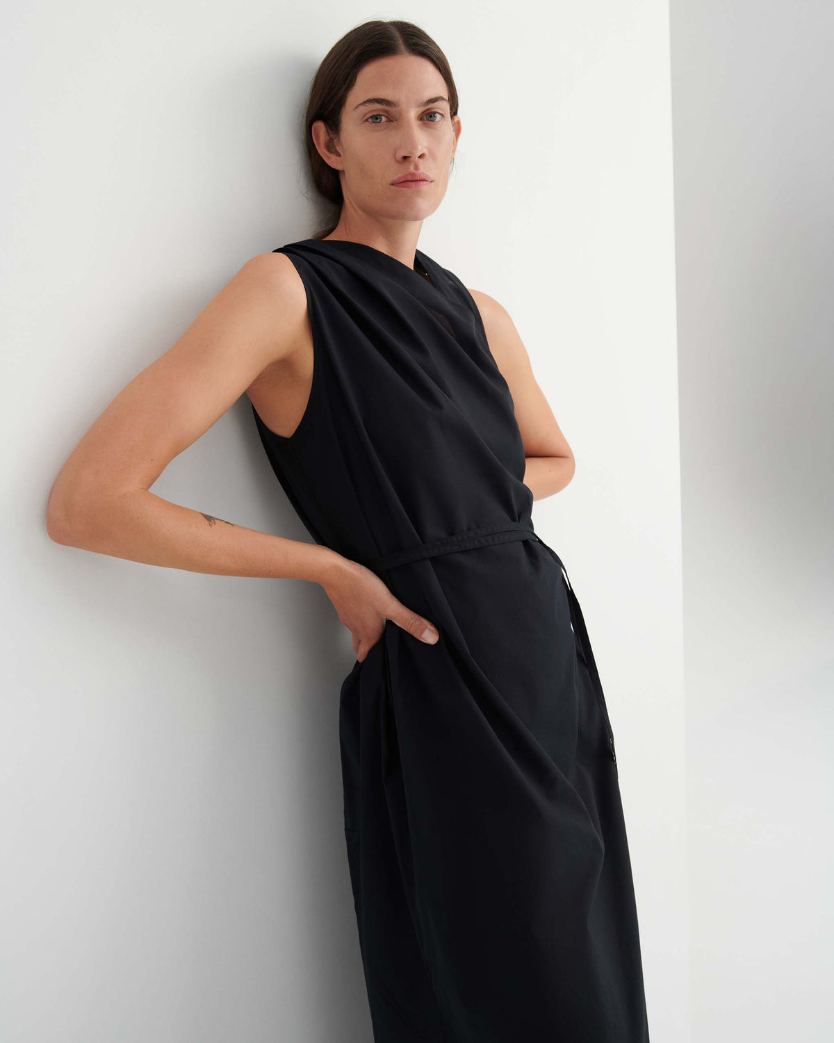 Person with long brown hair wearing a sleeveless black dress stands against a white wall with hands resting on hips, looking at the camera.