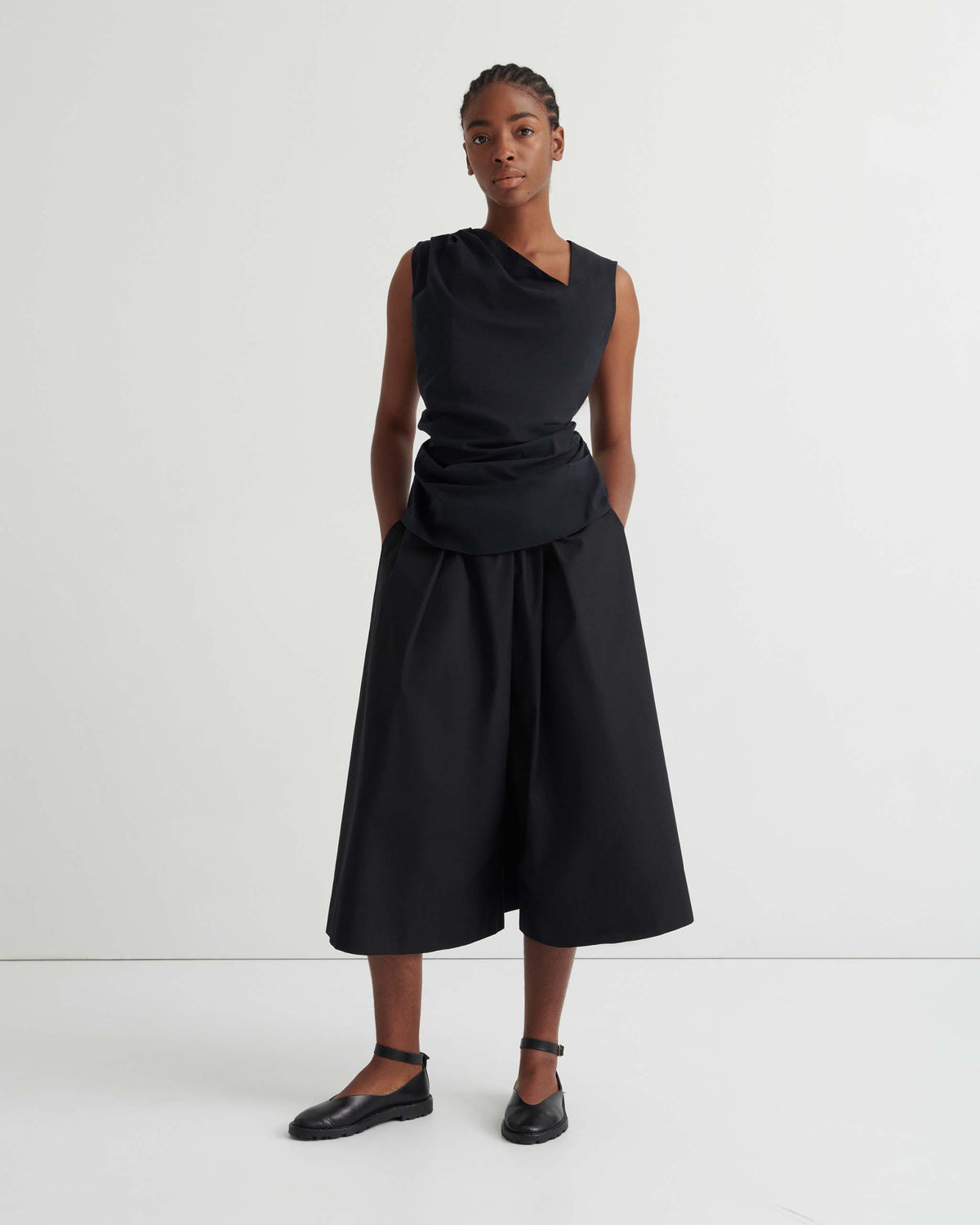 Person standing against a plain white background, wearing a sleeveless black top, wide black culottes, and black ankle-strap flats. Hands are in pockets, facing the camera.