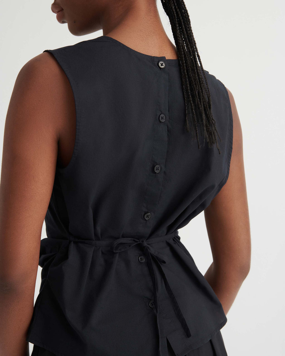 Person wearing a sleeveless black top with a buttoned back and a tied waist, shown from behind against a plain light background.