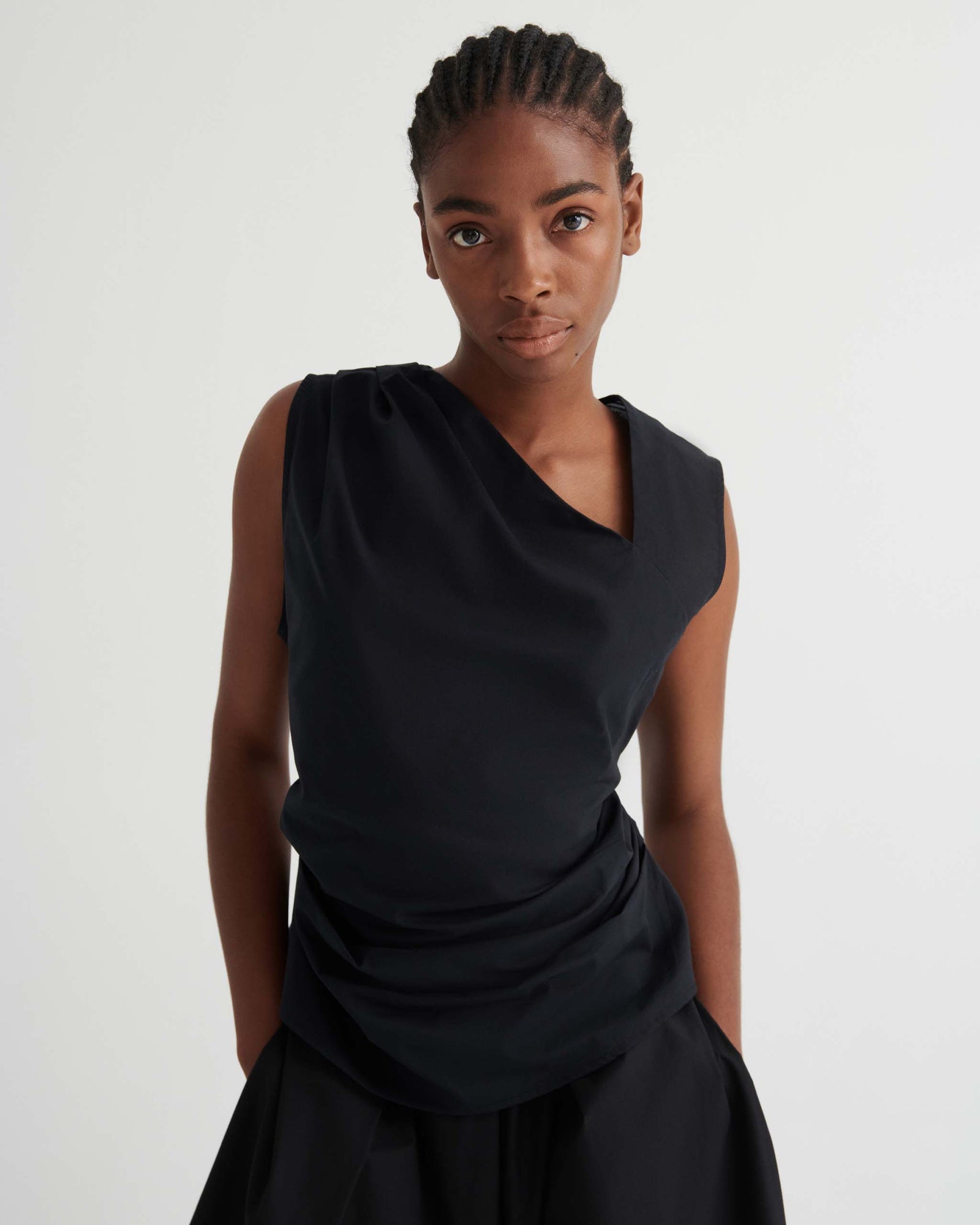 Person standing against a plain white background, wearing a sleeveless black draped top and a black skirt, looking directly at the camera with hands behind their back.