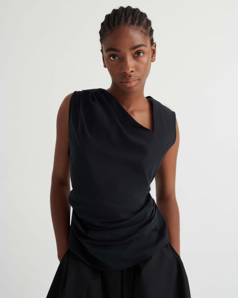 Person standing against a plain white background, wearing a sleeveless black draped top and a black skirt, looking directly at the camera with hands behind their back.