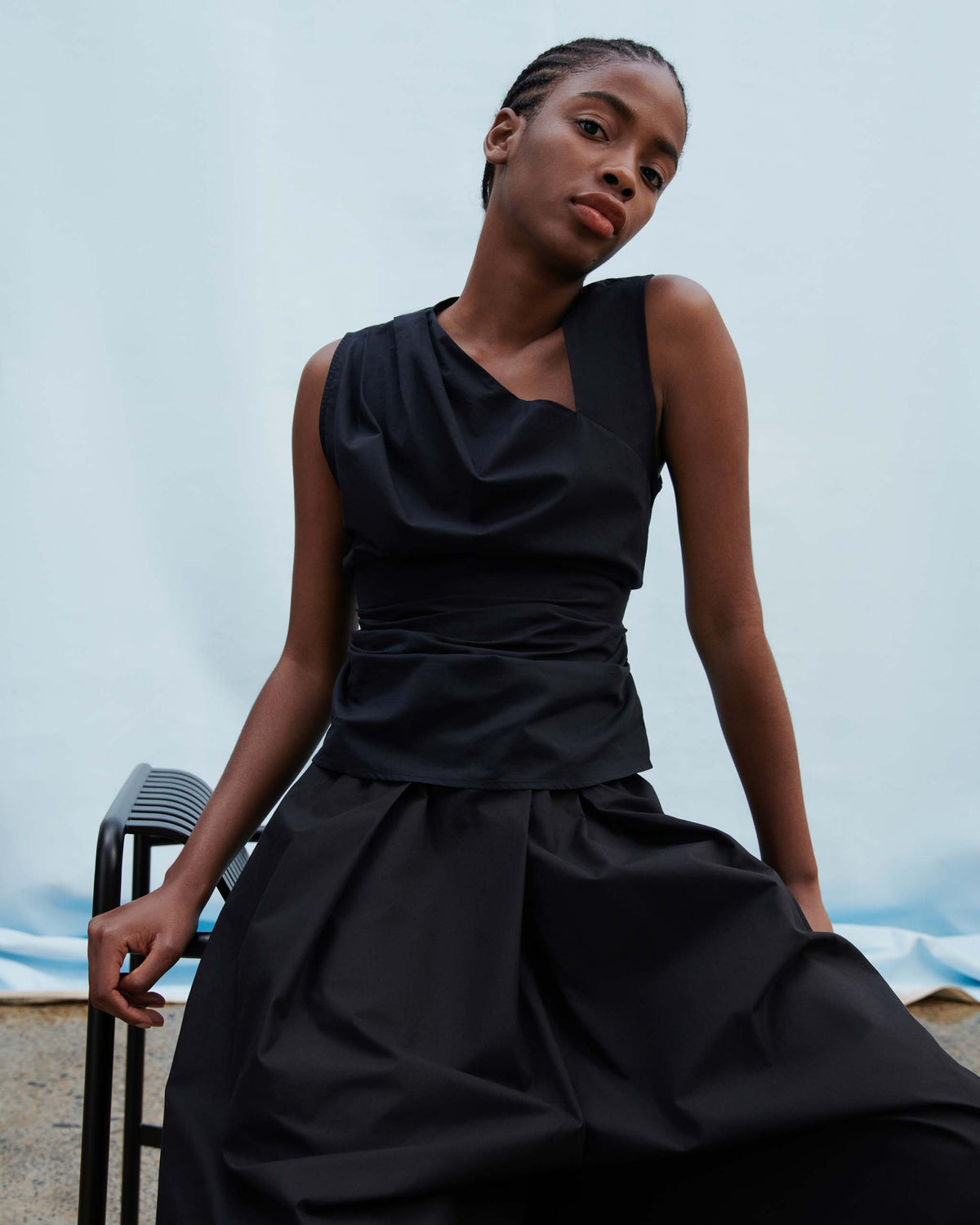 Person with braided hair wearing a sleeveless black dress sits on a black bench against a plain light blue backdrop, looking at the camera.