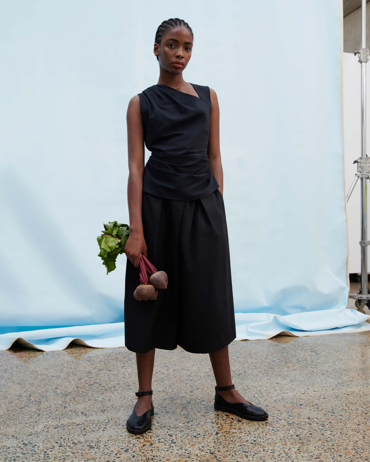 A person stands indoors on a speckled floor, wearing a sleeveless black top and culottes, holding beets in one hand. The background is a light blue draped cloth.