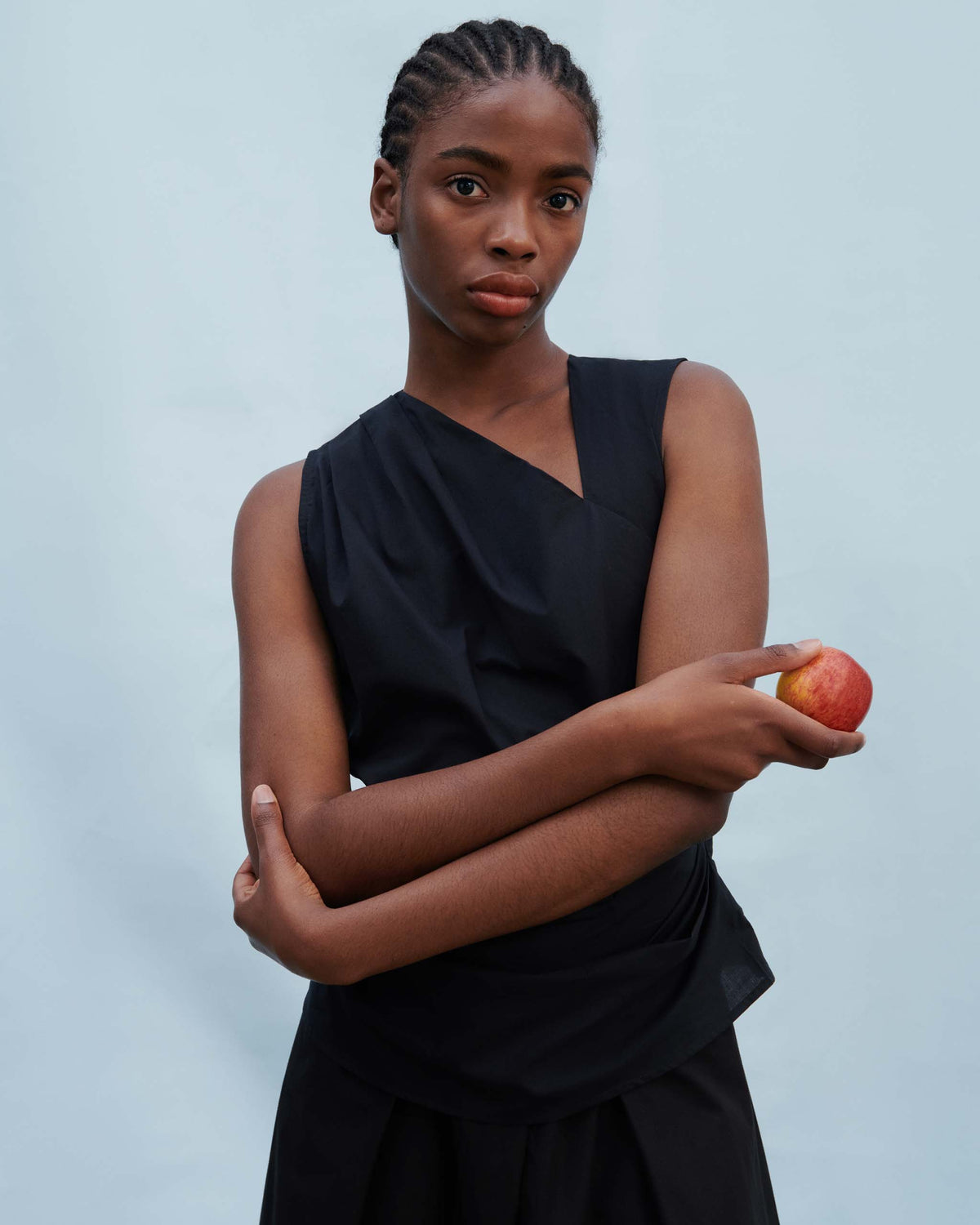 Person in a sleeveless black top stands against a plain light background, holding a red apple in one hand and looking at the camera.