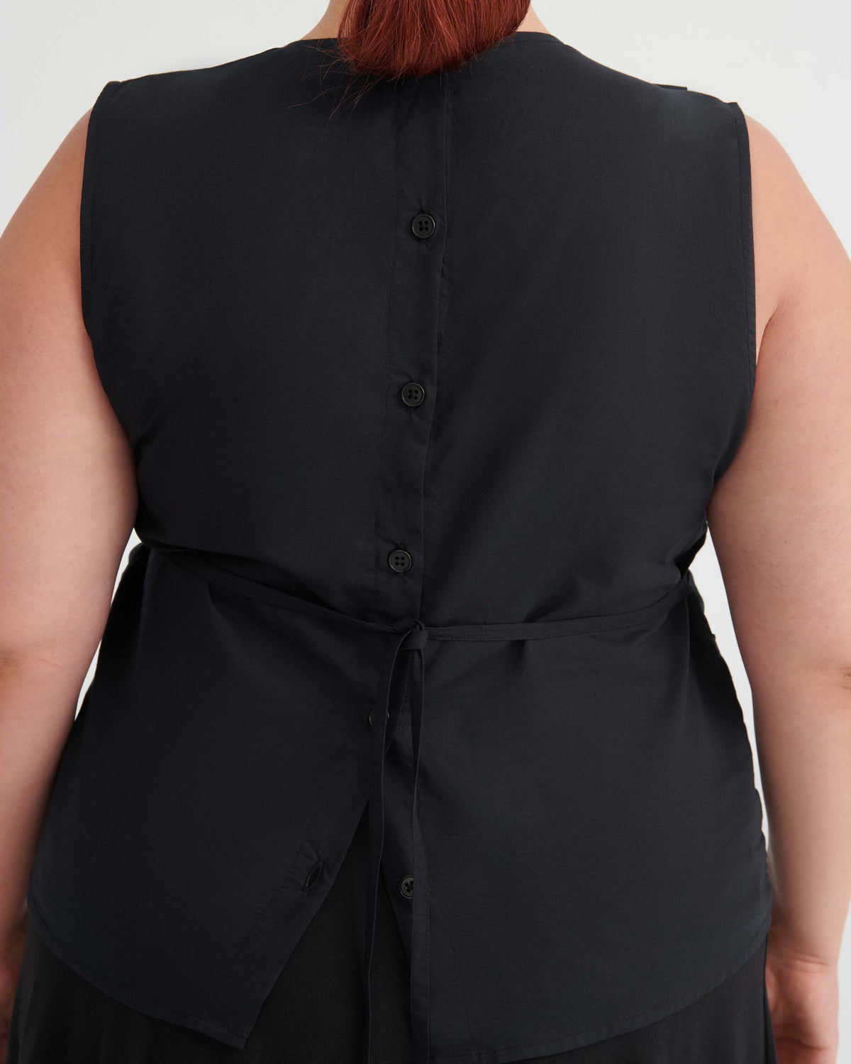 A person wearing a sleeveless black top seen from the back, featuring buttons down the center and a tie at the waist.
