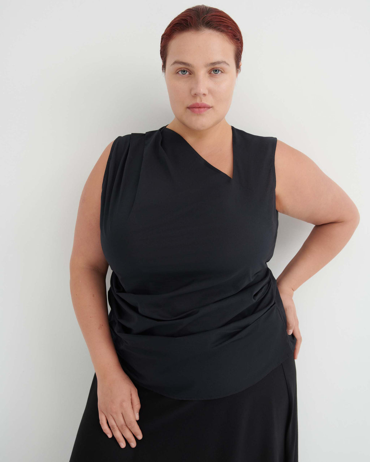 A woman with red hair tied back wears a sleeveless black top and black skirt, standing against a plain white background with one hand on her hip.