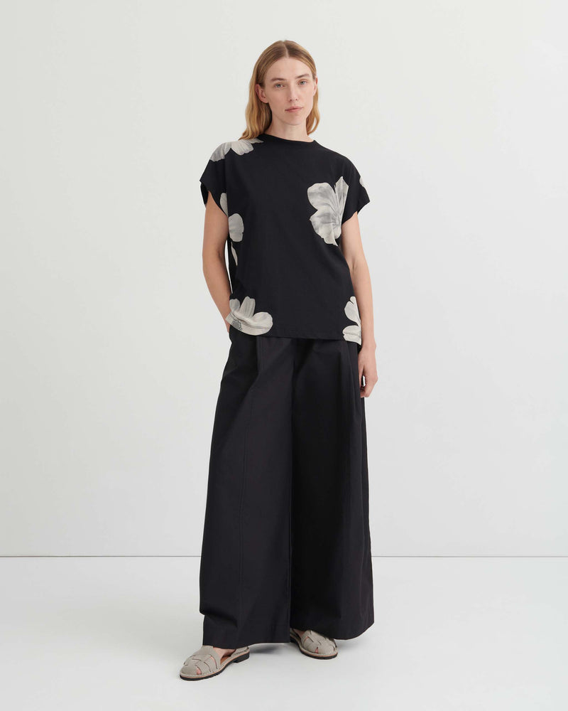 A person wears a black short-sleeved top with a white floral print, paired with wide-leg black trousers and grey sandals.