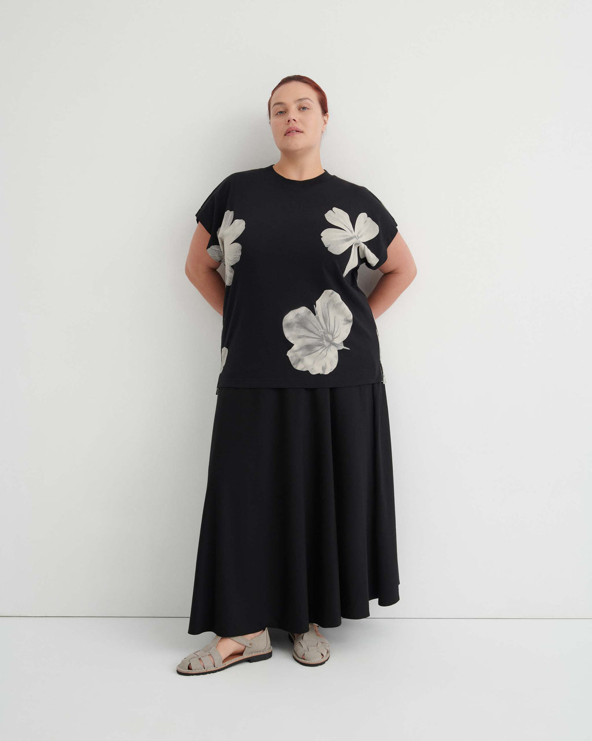 A person wearing a black t-shirt with a white floral print, a long black skirt, and light-colored sandals.