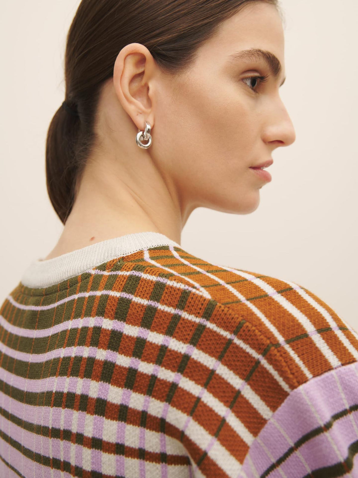 Side profile of a woman wearing an oversized Kowtow Gradient Knit Top plaid sweater and a silver hoop earring, with her hair styled in a low ponytail.
