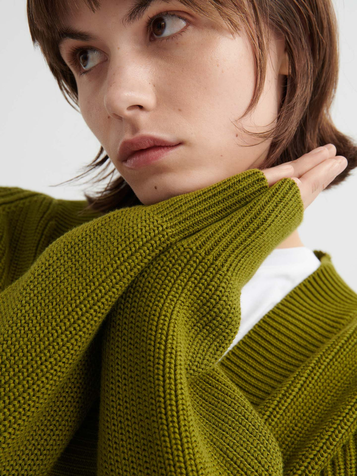 A person wearing the Hannes Cardigan – Field in green, featuring a relaxed fit, holds one hand to their face and gazes slightly away from the camera.
