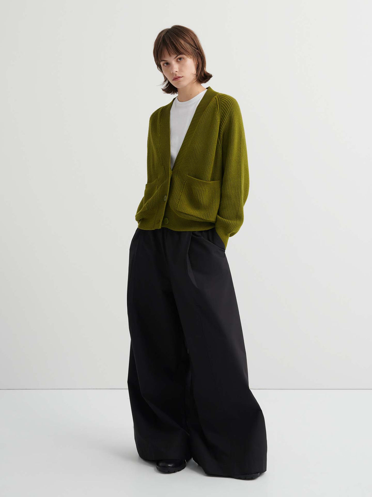 Model in a relaxed fit Hannes Cardigan – Field (olive green) over a white shirt with wide-leg black pants, posed against a plain white background—a modern update to women’s wardrobe essentials.