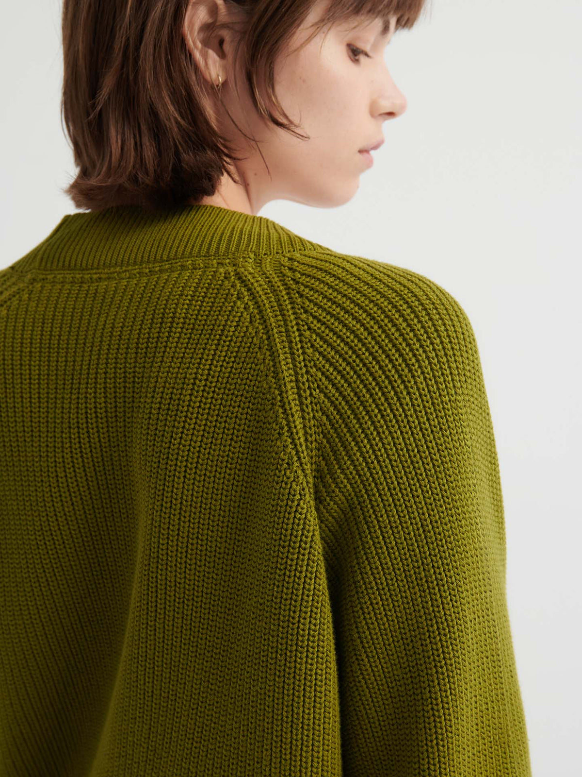 A person with short brown hair wears the Hannes Cardigan – Field, a relaxed fit olive-green knit, facing slightly away against a plain light background.