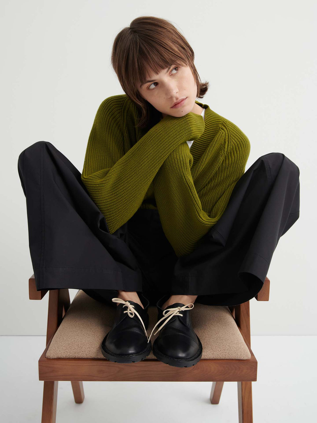 A person sits cross-legged on a chair, wearing a relaxed fit green Hannes Cardigan – Field, wide black pants, and black shoes with beige laces, looking to the side against a plain background. Women's clothing.