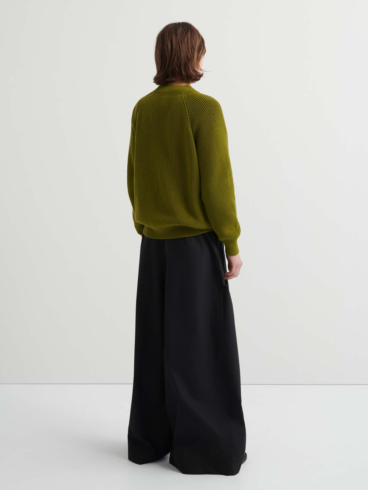 Person stands facing away in a relaxed fit Hannes Cardigan – Field (olive-green) and wide-leg black pants against a plain white background, showcasing an effortlessly stylish women's clothing look.