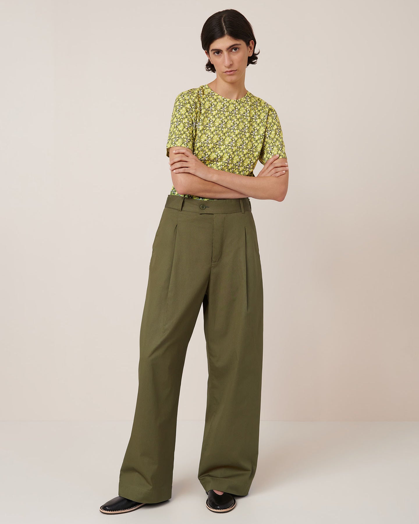 A person stands against a plain background wearing a yellow floral short-sleeve shirt tucked into Kowtow's Harmony Pant in Ivy, paired with black slip-on shoes. Refer to the Kowtow size guide for women's pant measurements.