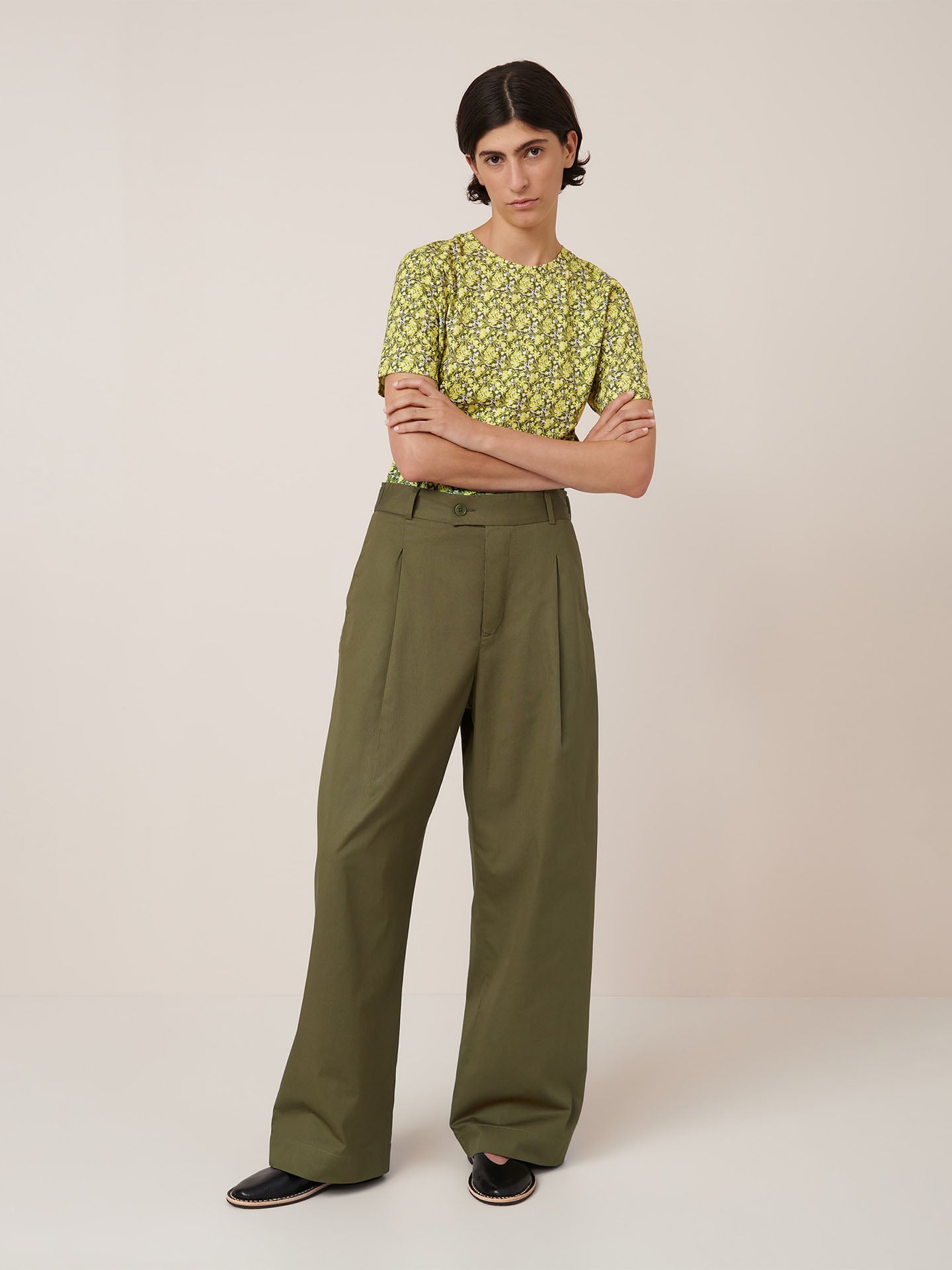 A person stands against a plain background wearing a yellow floral short-sleeve shirt tucked into Kowtow's Harmony Pant in Ivy, paired with black slip-on shoes. Refer to the Kowtow size guide for women's pant measurements.