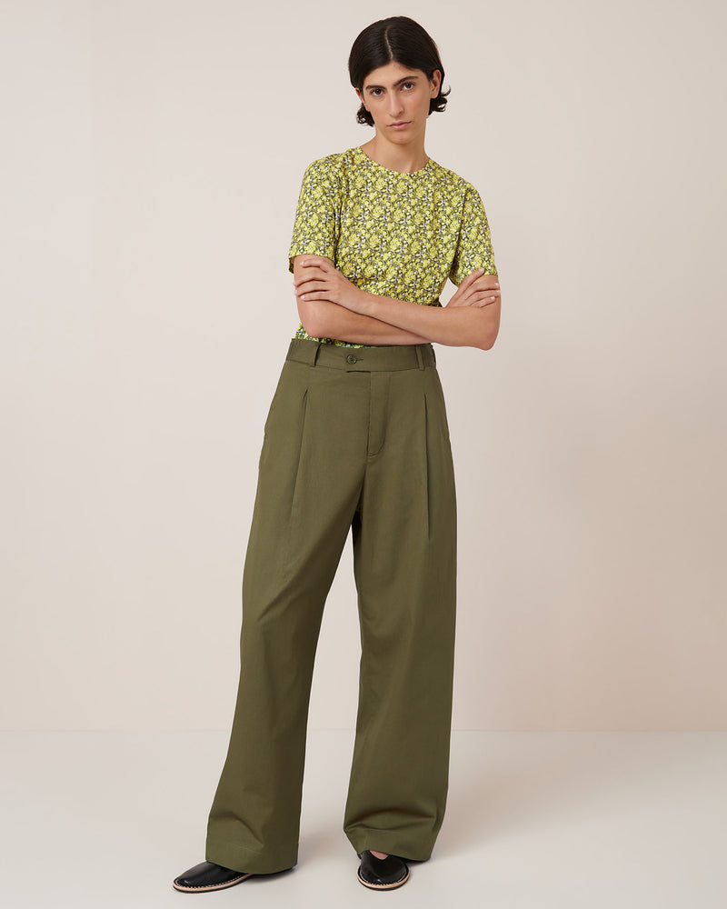 A person stands against a plain background wearing a yellow floral short-sleeve shirt tucked into Kowtow's Harmony Pant in Ivy, paired with black slip-on shoes. Refer to the Kowtow size guide for women's pant measurements.