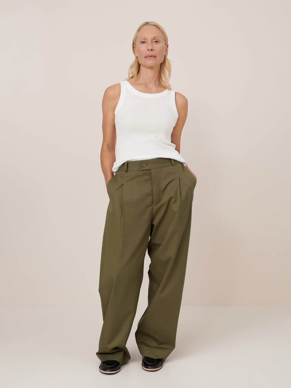 A person stands against a plain background, wearing a white sleeveless top, Kowtow’s Harmony Pant – Ivy in olive green, and black shoes with hands in pockets.