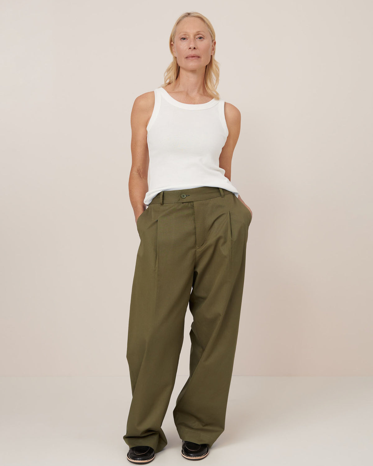 A person stands against a plain background, wearing a white sleeveless top, Kowtow’s Harmony Pant – Ivy in olive green, and black shoes with hands in pockets.