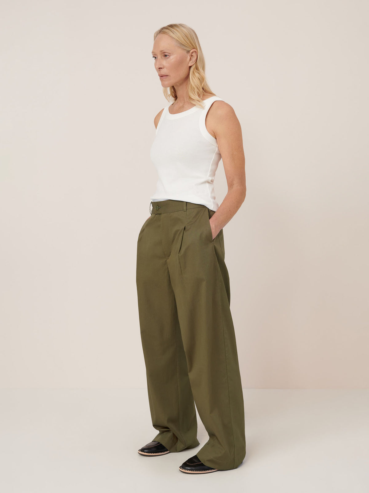 A woman stands against a plain background wearing a white sleeveless top, black shoes, and Kowtow’s Harmony Pant – Ivy in a relaxed mid-rise fit.