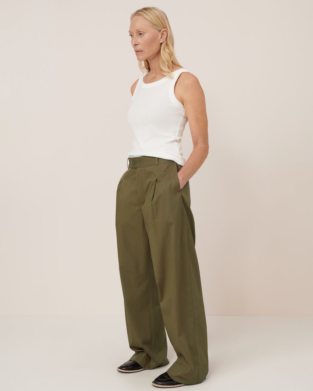 A woman stands against a plain background wearing a white sleeveless top, black shoes, and Kowtow’s Harmony Pant – Ivy in a relaxed mid-rise fit.