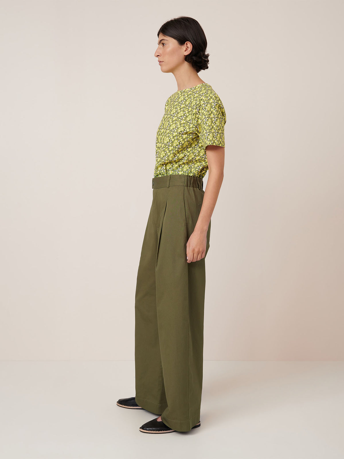 A person stands sideways against a plain background, wearing a green floral top, Kowtow Harmony Pant – Ivy high-waisted wide-leg pants with a relaxed fit, and black slip-on shoes.