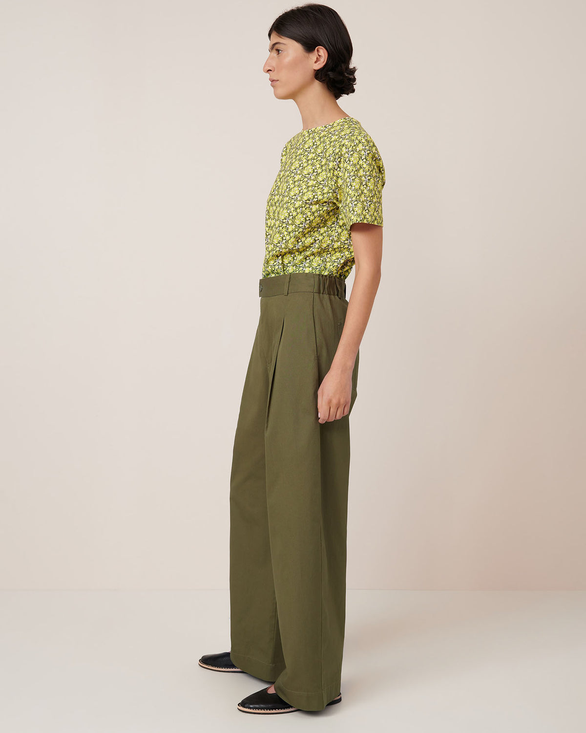 A person stands sideways against a plain background, wearing a green floral top, Kowtow Harmony Pant – Ivy high-waisted wide-leg pants with a relaxed fit, and black slip-on shoes.