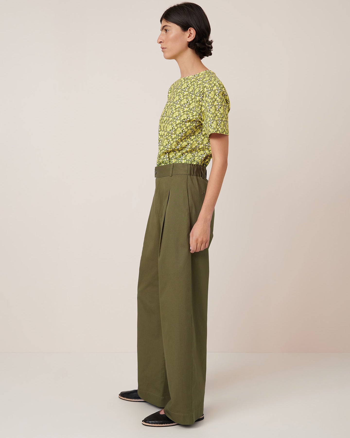 A person stands against a plain background wearing a yellow floral short-sleeve shirt tucked into Kowtow's Harmony Pant in Ivy, paired with black slip-on shoes. Refer to the Kowtow size guide for women's pant measurements.