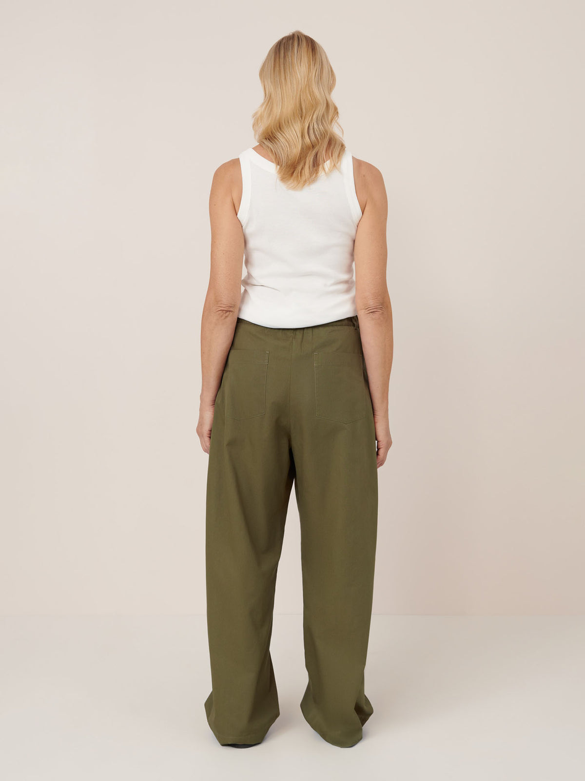 A person with shoulder-length blonde hair, wearing a white sleeveless top and mid-rise relaxed fit Harmony Pant – Ivy by Kowtow, stands facing away from the camera against a plain background to illustrate women&#39;s pant measurements for the size guide.