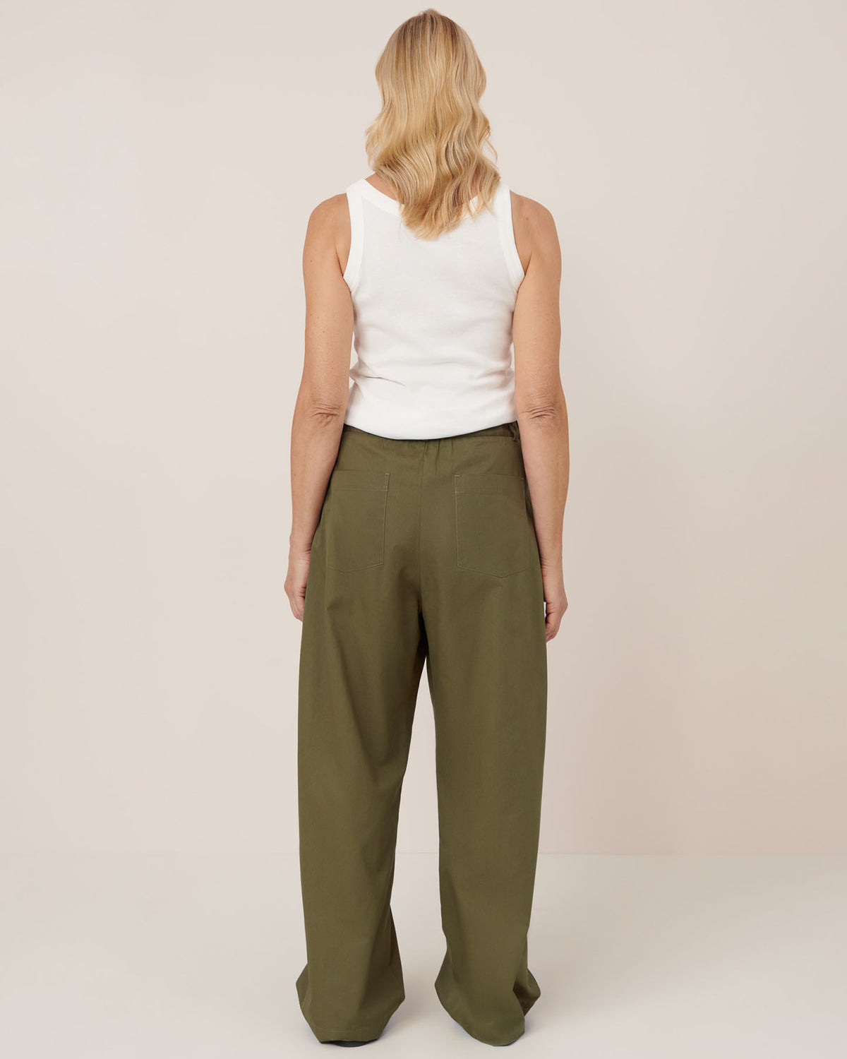 A person with shoulder-length blonde hair, wearing a white sleeveless top and mid-rise relaxed fit Harmony Pant – Ivy by Kowtow, stands facing away from the camera against a plain background to illustrate women's pant measurements for the size guide.