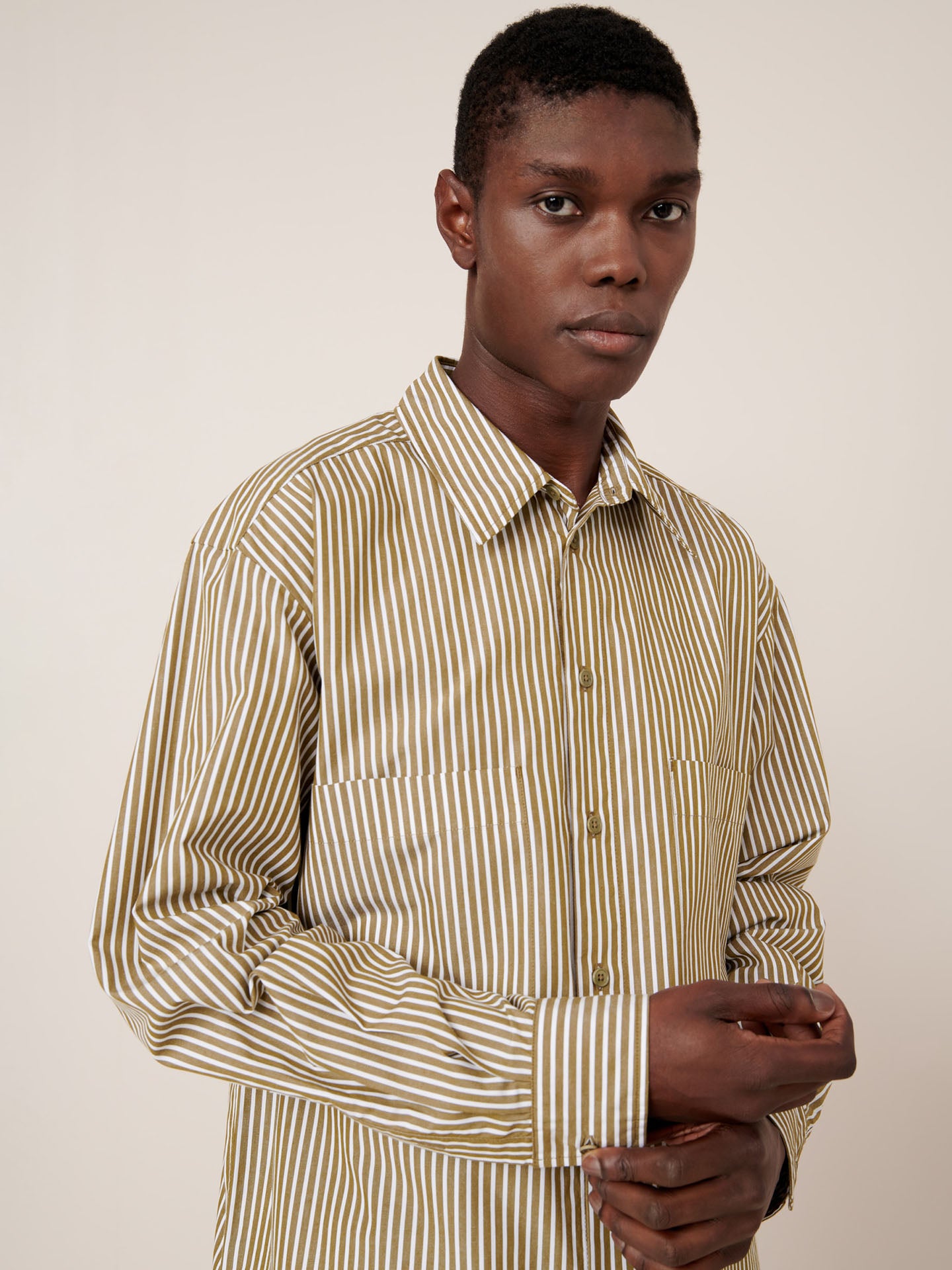 A person wearing the Kowtow Henry Shirt in Treetop Stripe, featuring beige and white vertical stripes and a relaxed fit, stands against a plain light background, looking at the camera.