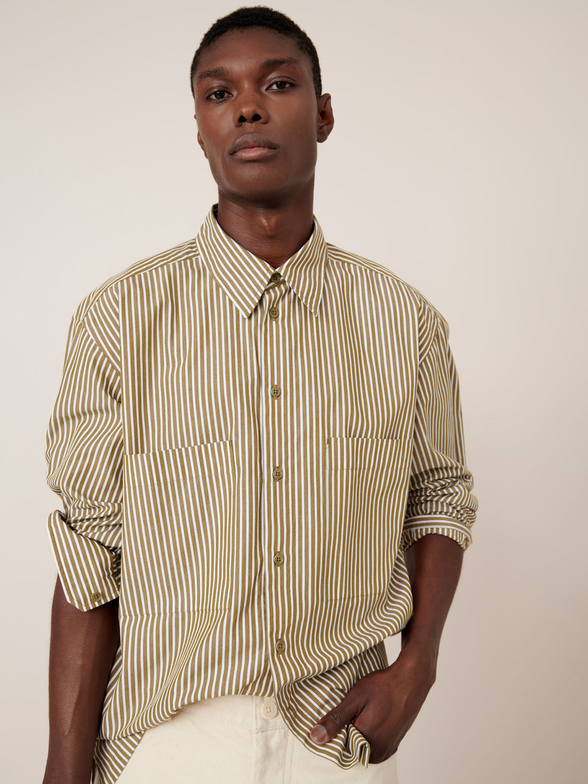 A person wearing the Henry Shirt – Treetop Stripe by Kowtow, featuring beige and olive vertical stripes with rolled sleeves and a relaxed fit, stands against a plain light background.