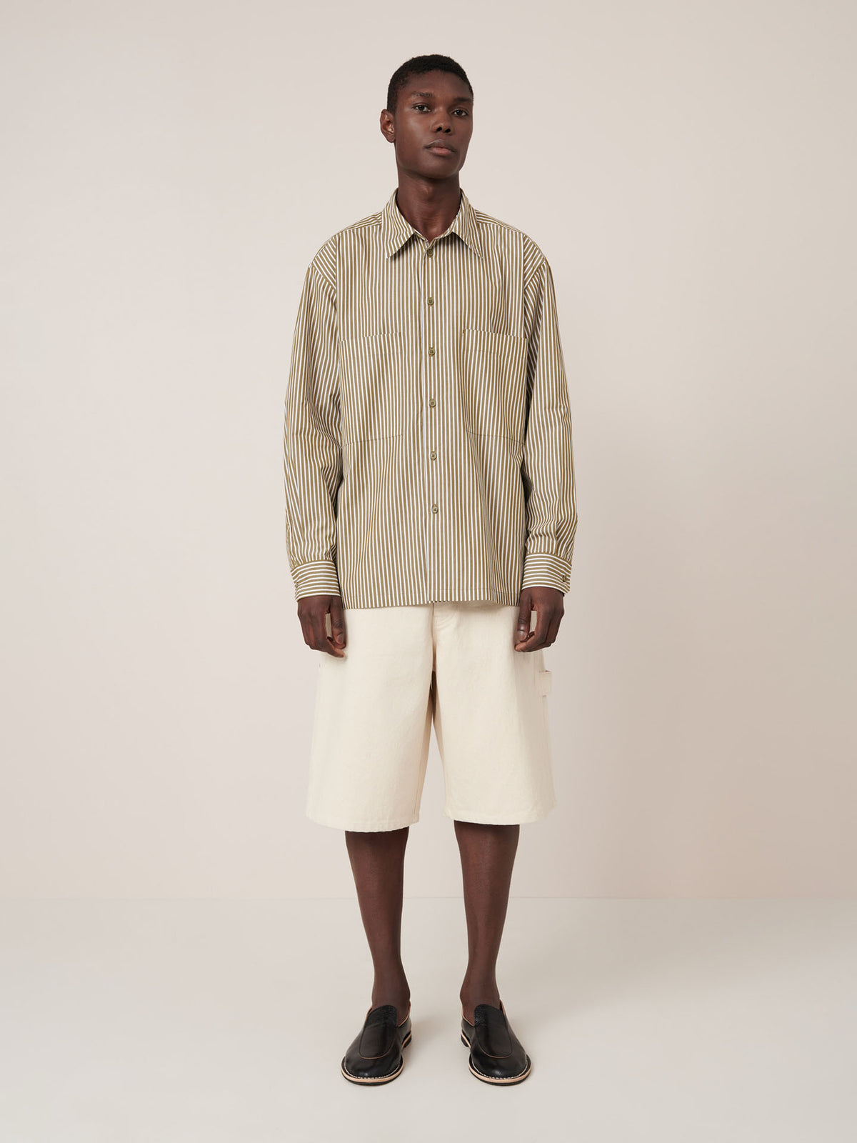 A man stands facing forward against a plain light background, wearing the Kowtow Henry Shirt in Treetop Stripe and cream knee-length shorts, paired with black slip-on shoes.