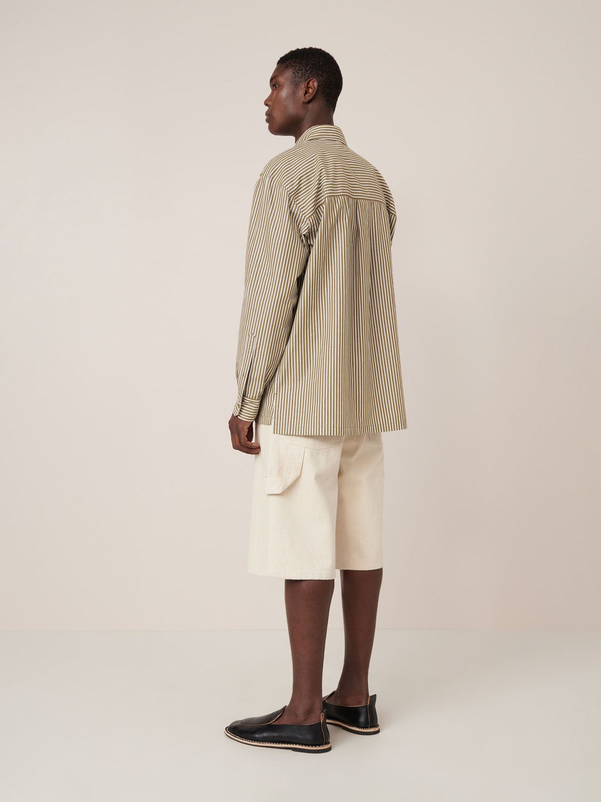 A person stands facing away, wearing the Kowtow Henry Shirt in Treetop Stripe—a relaxed-fit, beige and brown striped long-sleeve—paired with cream knee-length shorts with pockets and black slip-on shoes against a plain background.