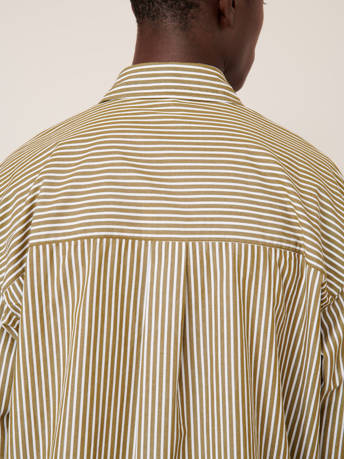 A person is seen from the back wearing Kowtow’s Henry Shirt – Treetop Stripe, which features olive and white horizontal and vertical stripes in a relaxed fit.