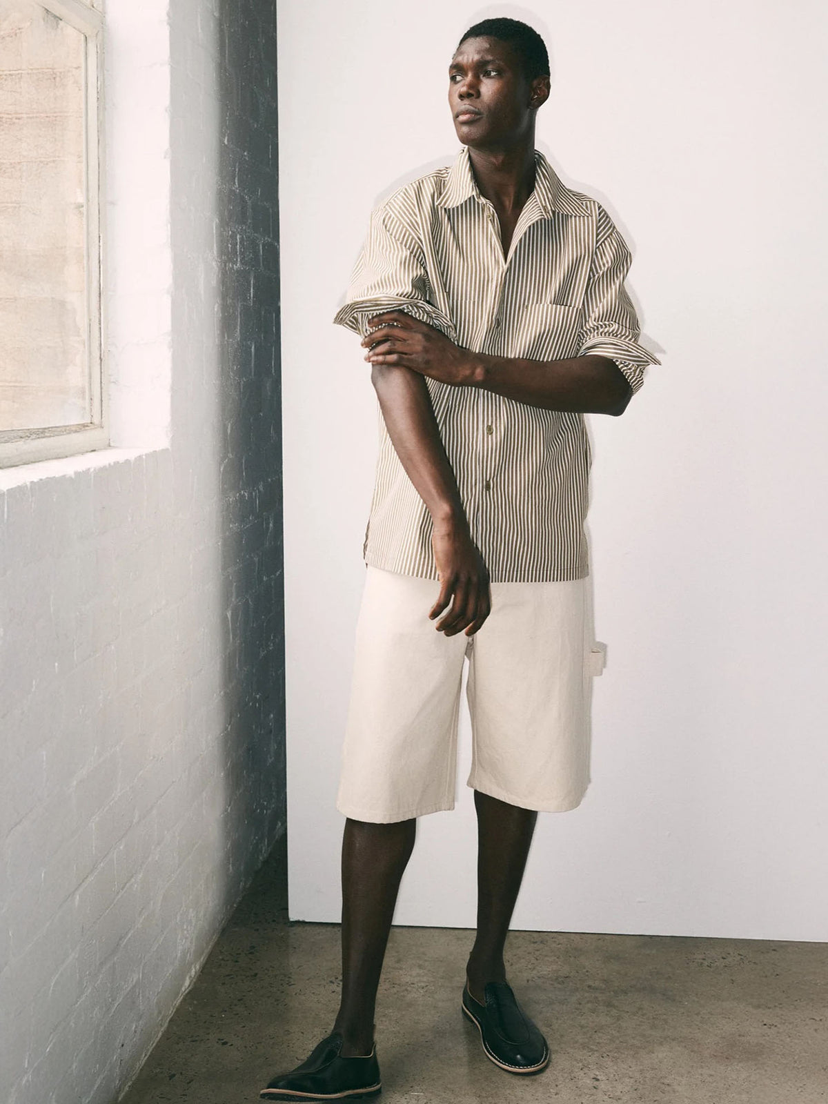A man stands indoors near a window, wearing the Kowtow Henry Shirt in Treetop Stripe, light knee-length relaxed-fit shorts, and black slip-on shoes.