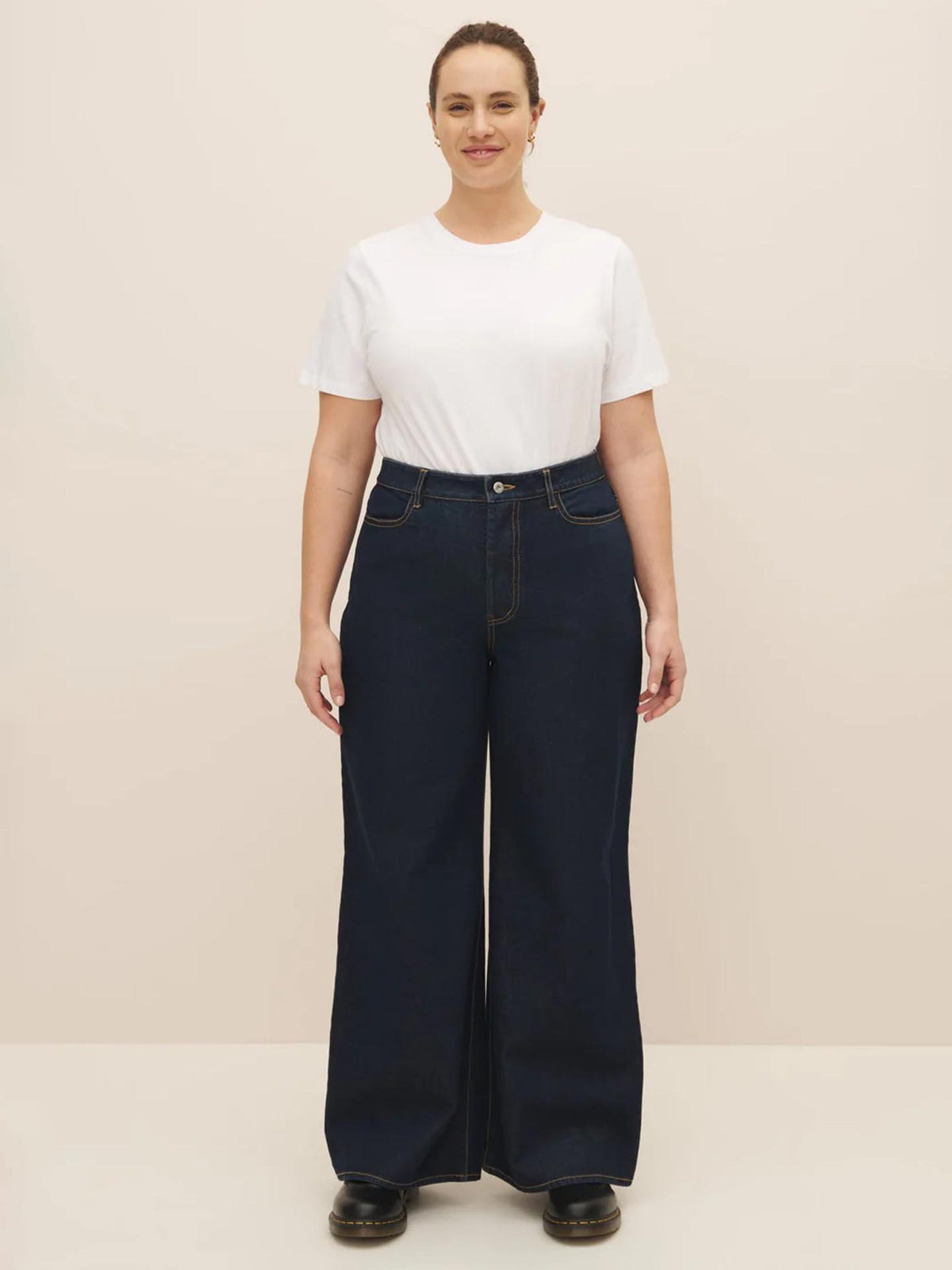 Woman standing in a neutral pose wearing a white t-shirt and Kowtow High Puddle Jeans – Indigo Denim against a soft beige background.