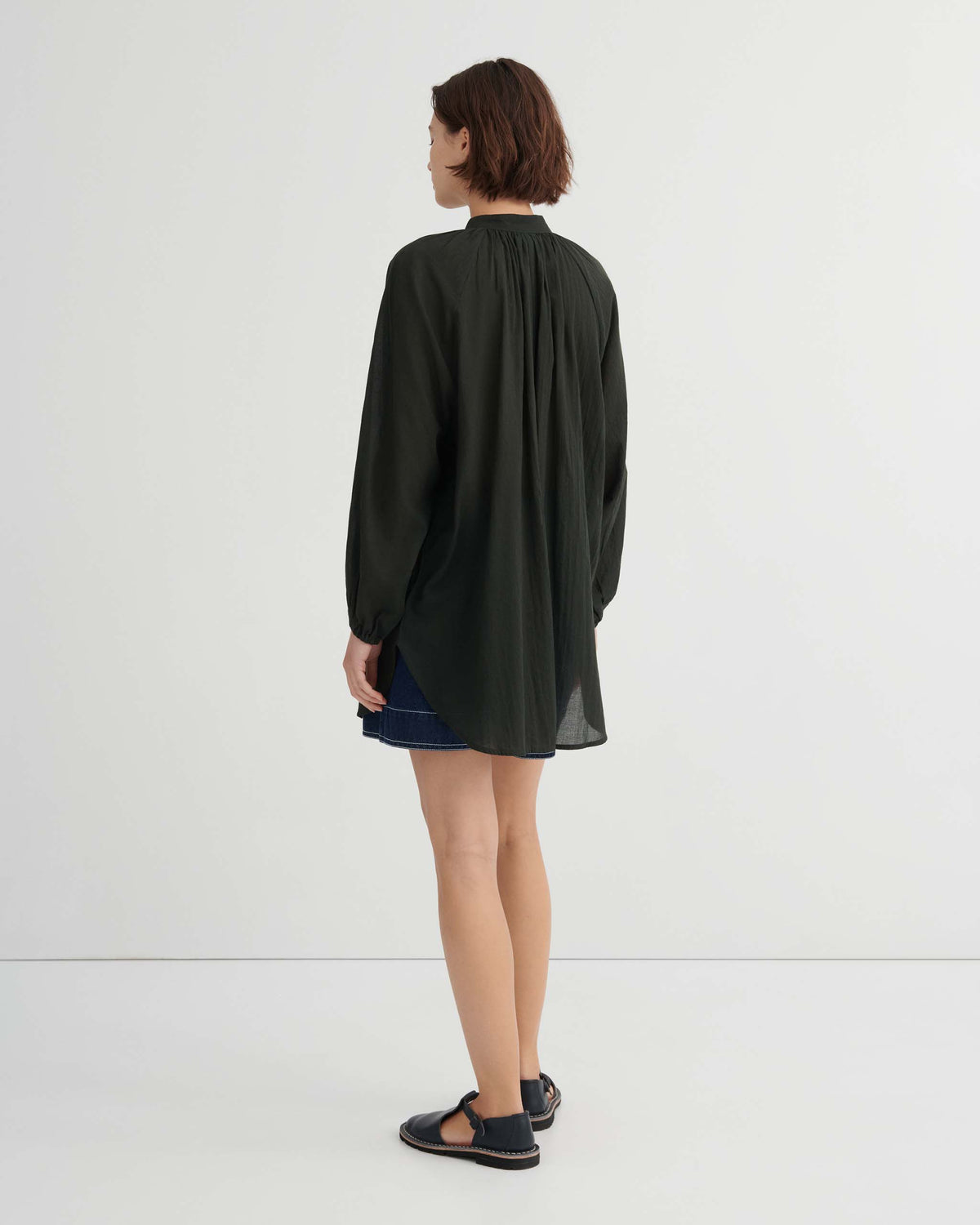 A person seen from behind wearing an oversized dark green long-sleeved shirt and a denim mini skirt with dark sandals.